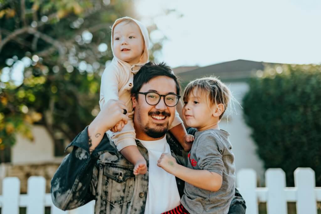 Fathers holds two children, one in his left arm and one on his right shoulder