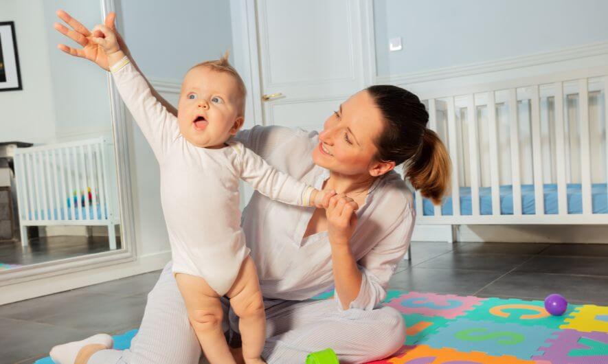 Woman sitting on the floor of a nursery on colorful playmat holding the hands of an excited toddler, standing next to her with an animated facial expression