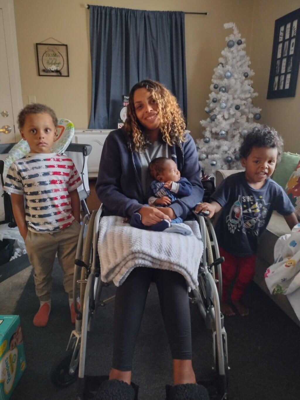 HFA Parent Graduate of the Year sits, center frame, in her wheelchair, flanked by two of her children, each standing with their heads about to her shoulders. She is holding her youngest child, an infant, on her lap.