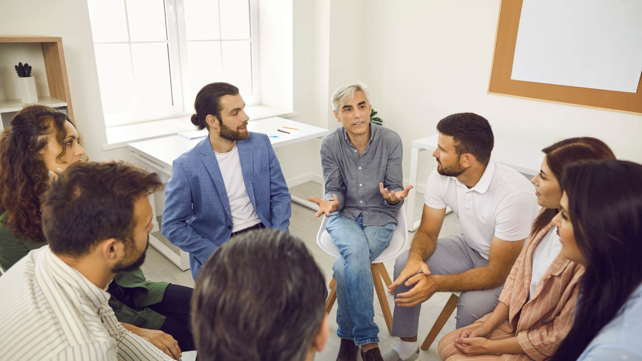 Group sits in a circle, listening to individual at center of frame. They speak, legs crossed, hands open in conversation.