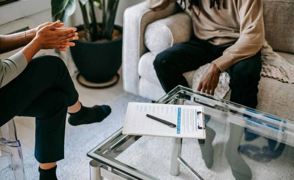 A clipboard sits on a table between two people, with only their legs displayed. One sits n a couch and the other in a nearby chair.