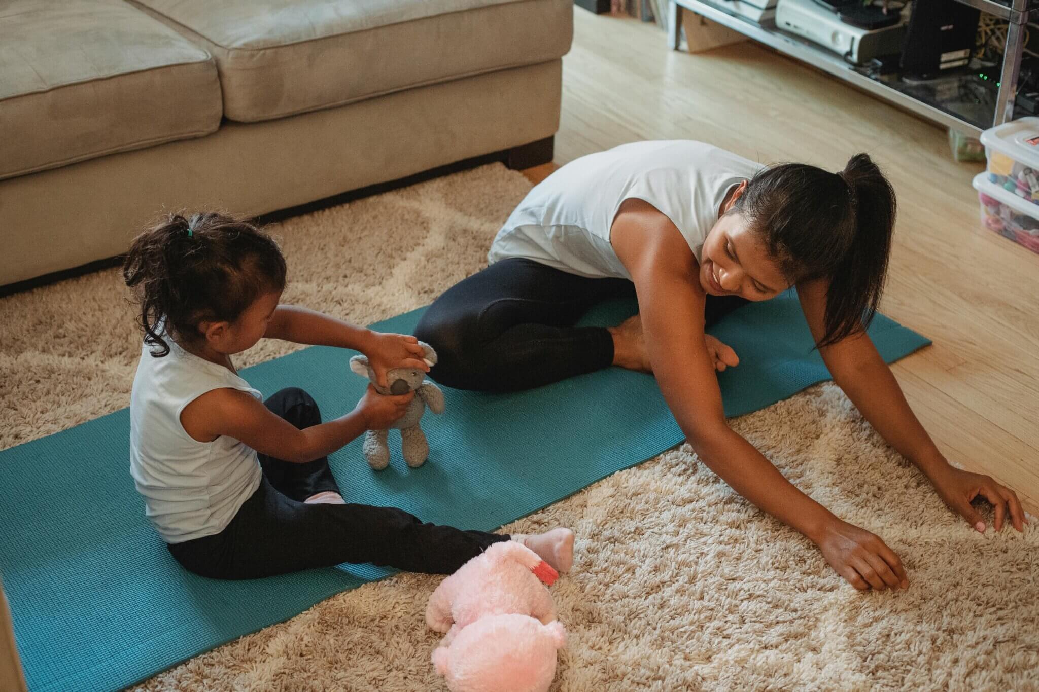 A mom sits on the floor on a yoga mat, with her daugher sitting next to her, both stretching.