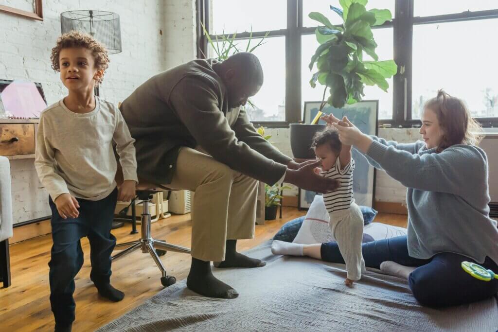 Active family of four, 2 parents and 2 children play in the living room. The baby tries to stand between the two parents.