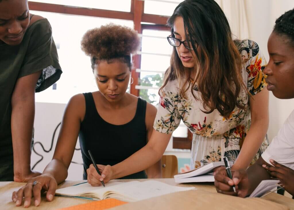 Team looks down at shared notebook, one member, standing, leans in to write on the paper with a pen.