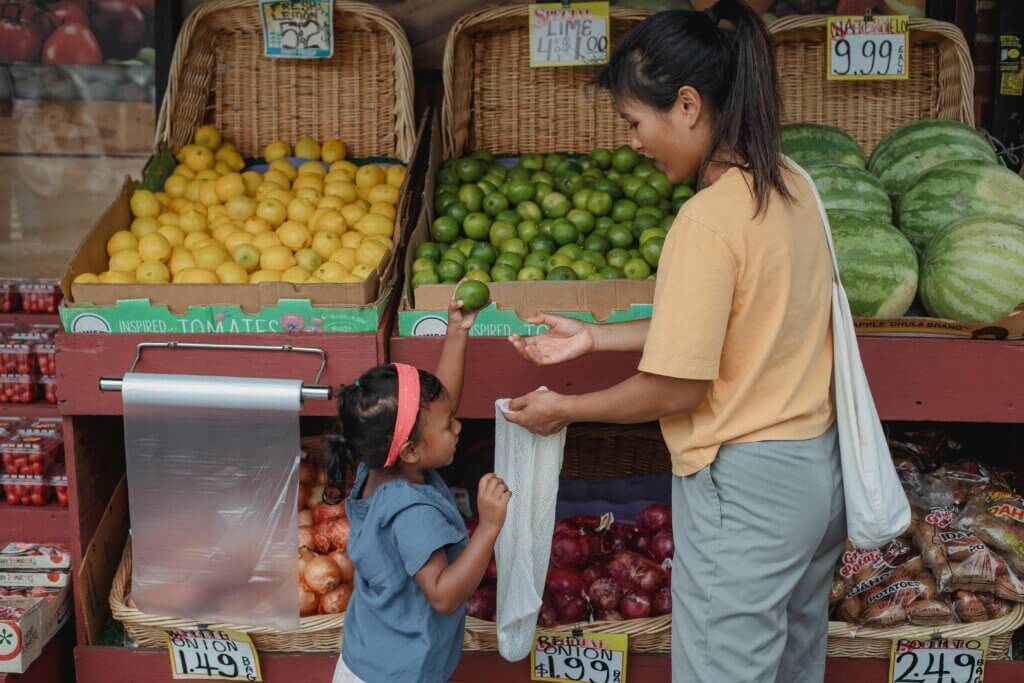 Mom and daughter stand next to a big, multi-tiered fruit stand, adding fresh fruit to their bag.