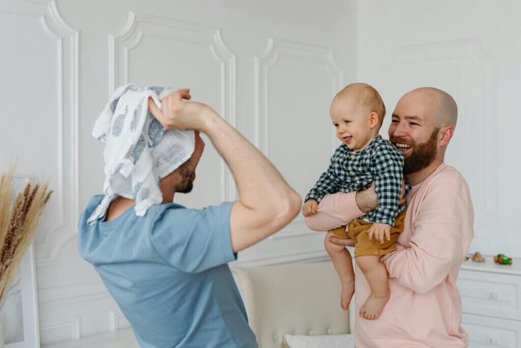 While one parent holds their child, facing the other, who acts silly with clothes on their head