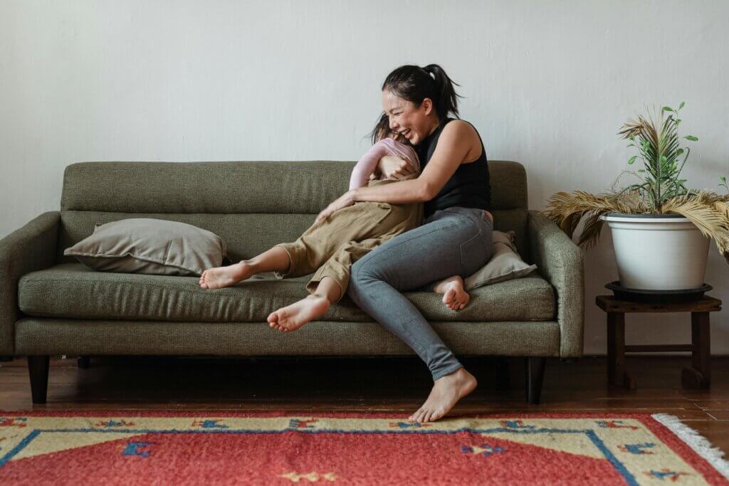 Mom sitting on a couch, facing sideways, with one foot curled undernearth her and another dangling off the side, tightly embraces her child